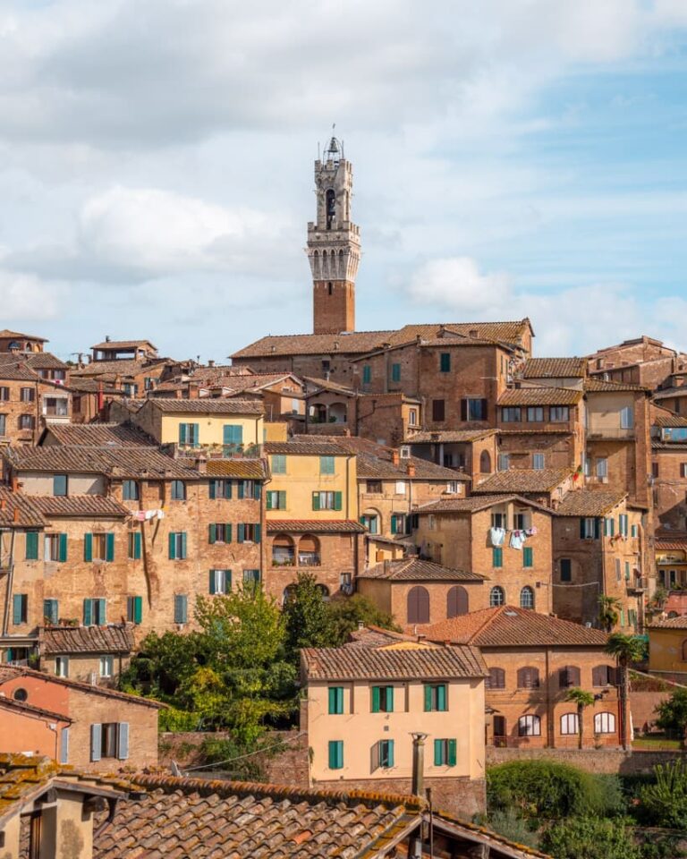 Toppen af Torre del Mangia bag huse på bakke i Siena